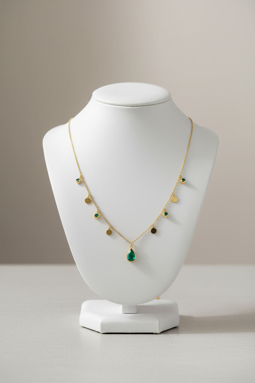 A delicate gold chain necklace featuring a teardrop-shaped green gemstone pendant at the center, with small gold disc charms and a few tiny green stone accents along the chain, displayed on a clean white background.