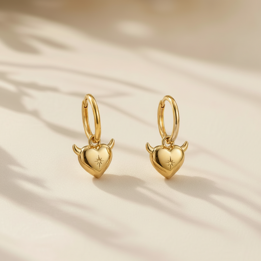 A pair of small gold hoop earrings with heart-shaped dangling pendants featuring tiny horns and a star detail, photographed on a warm beige surface with soft leaf shadows.