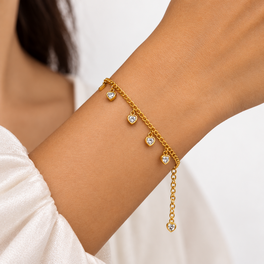 A gold chain bracelet with three small heart charms, worn on a person's wrist. The bracelet features clear rhinestones in the center of each heart.