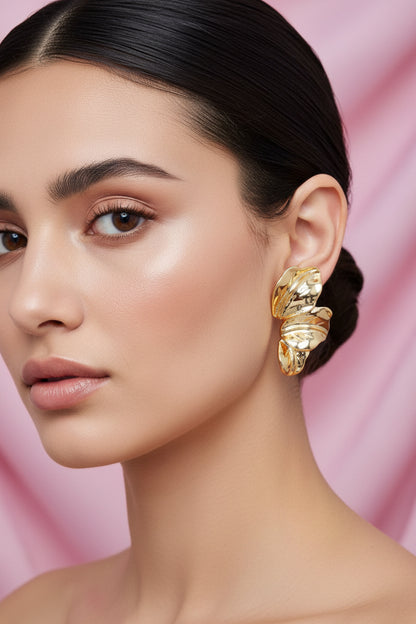 Gold leaf-shaped earrings on a white card with text, against a pink background