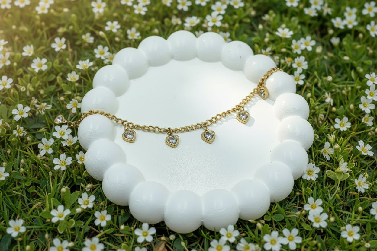 A delicate gold chain bracelet with four small heart charms, each set with a clear crystal, displayed on a white beaded jewelry stand against a pink satin background.