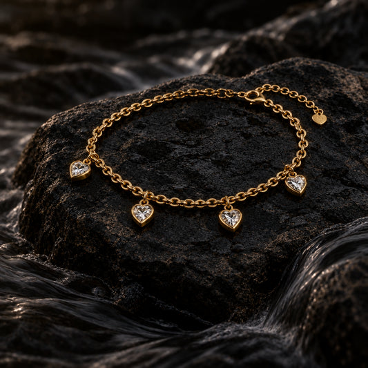 A delicate gold chain bracelet with four small heart charms, each set with a clear crystal, displayed on a white beaded jewelry stand against a pink satin background.