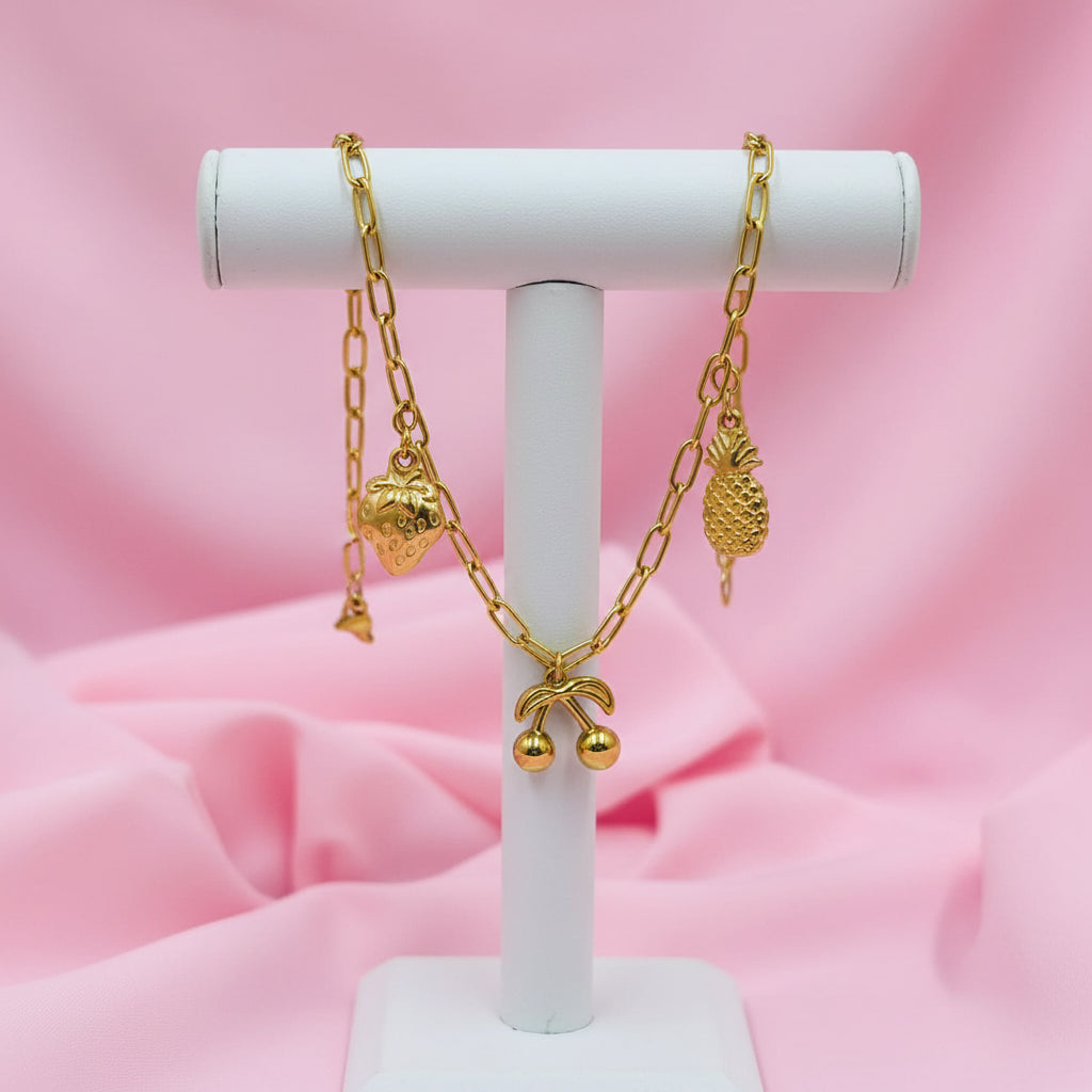 A gold chain bracelet with small charms, including a pineapple, cherries, and a strawberry, displayed on a white beaded bracelet over a pink satin background.