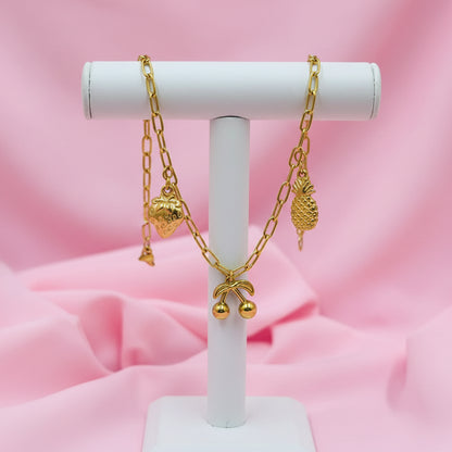 A gold chain bracelet with small charms, including a pineapple, cherries, and a strawberry, displayed on a white beaded bracelet over a pink satin background.