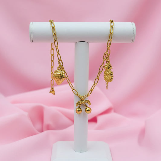 A gold chain bracelet with small charms, including a pineapple, cherries, and a strawberry, displayed on a white beaded bracelet over a pink satin background.