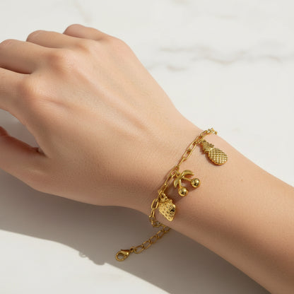 A gold chain bracelet with small charms, including a pineapple, cherries, and a strawberry, displayed on a white beaded bracelet over a pink satin background.