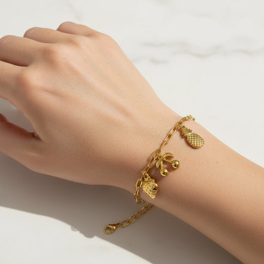 A gold chain bracelet with small charms, including a pineapple, cherries, and a strawberry, displayed on a white beaded bracelet over a pink satin background.