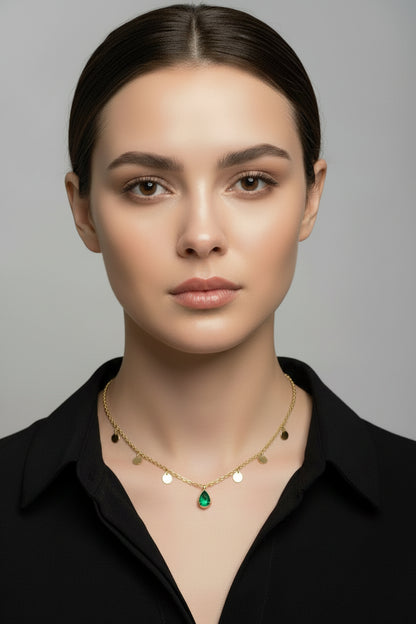 A delicate gold necklace with small gold beads and a central green teardrop pendant, displayed on a white jewelry bust with a wooden stand against a brown wood-paneled background.