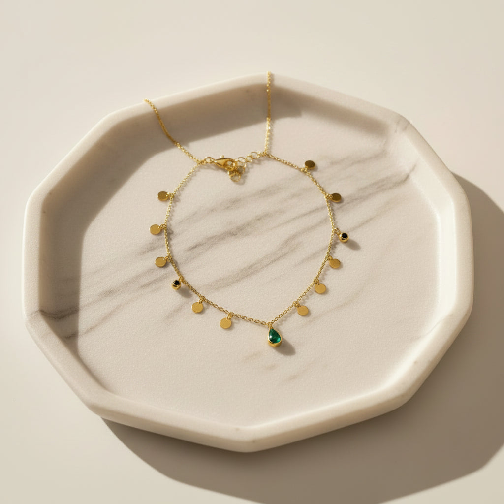 A delicate gold necklace with small gold beads and a central green teardrop pendant, displayed on a white jewelry bust with a wooden stand against a brown wood-paneled background.