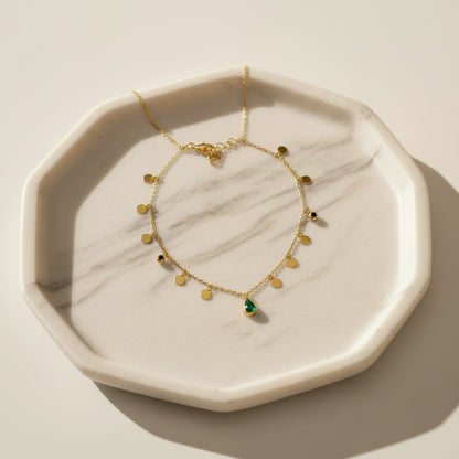 A delicate gold necklace with small gold beads and a central green teardrop pendant, displayed on a white jewelry bust with a wooden stand against a brown wood-paneled background.