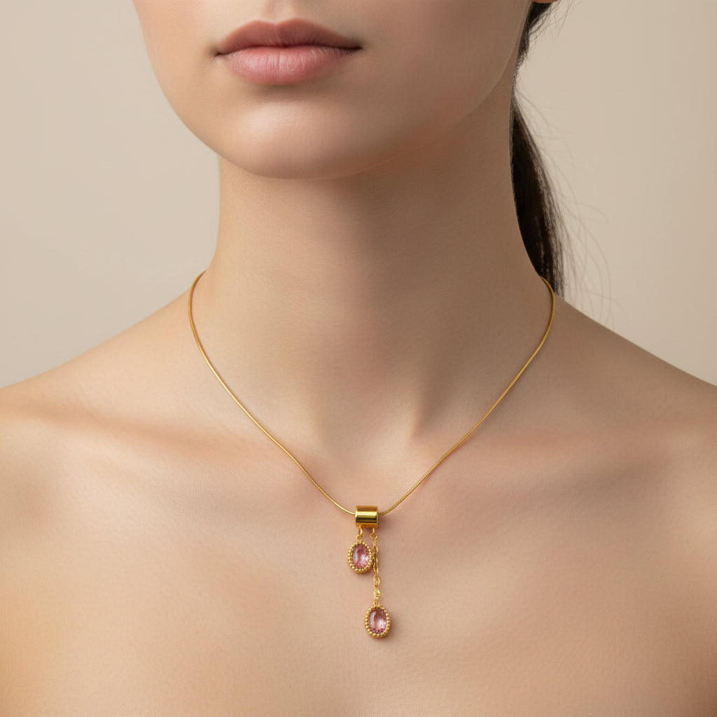 A gold chain necklace with two dangling oval pink gemstone pendants, displayed on a white beaded tray with pink fabric around it.
