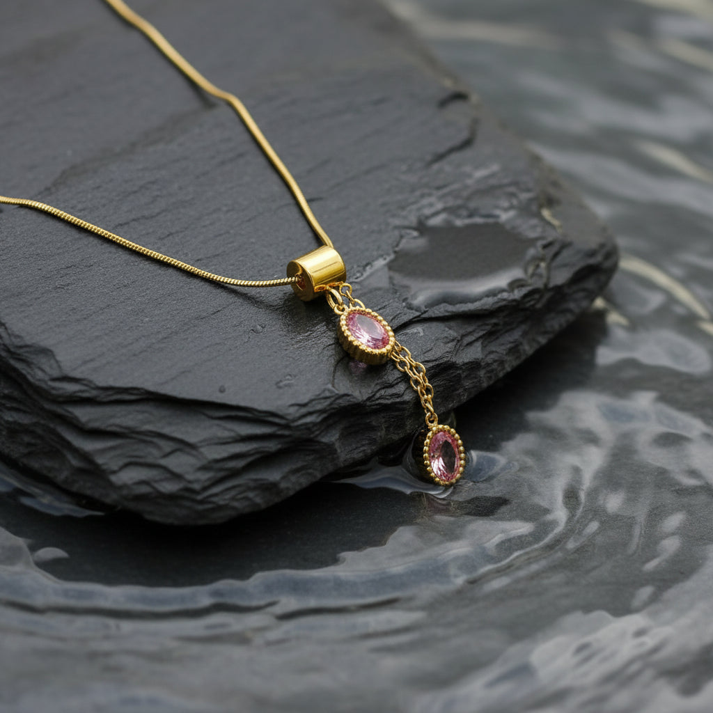A gold chain necklace with two dangling oval pink gemstone pendants, displayed on a white beaded tray with pink fabric around it.