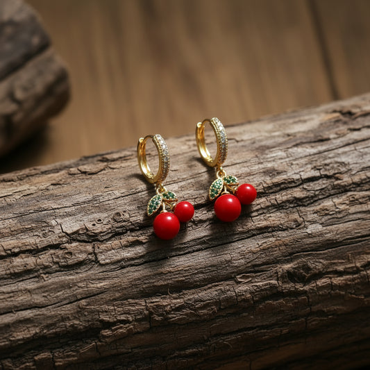 A pair of gold huggie hoop earrings with red bead drops and green leaf accents, displayed on a white card labeled 'Delicate' against a pink and white background.