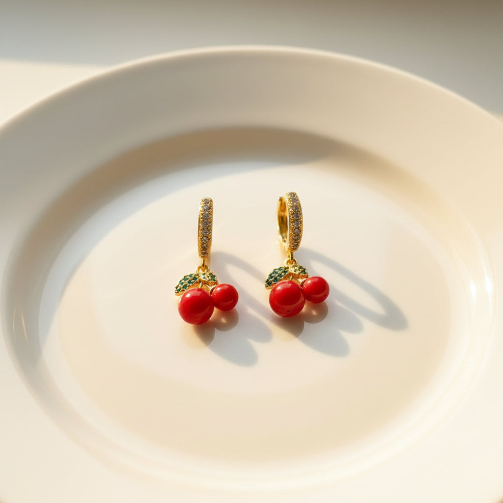 A pair of gold huggie hoop earrings with red bead drops and green leaf accents, displayed on a white card labeled 'Delicate' against a pink and white background.