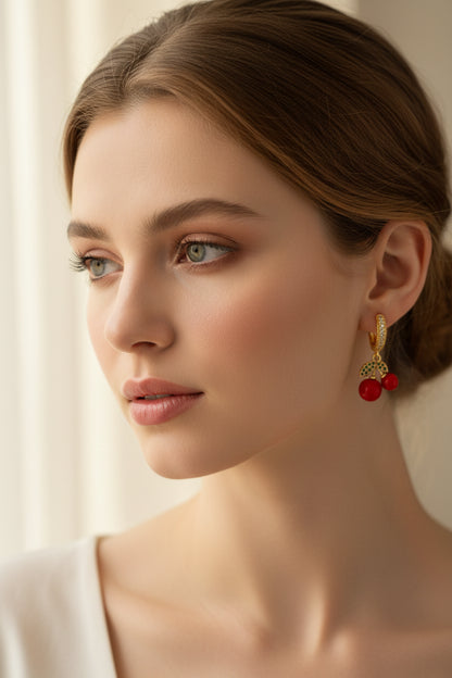 A pair of gold huggie hoop earrings with red bead drops and green leaf accents, displayed on a white card labeled 'Delicate' against a pink and white background.