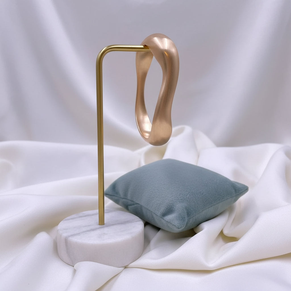 A polished gold-colored curved bangle bracelet displayed on a small dusty blue velvet cushion, set against a background of draped white fabric.