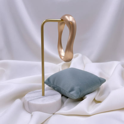 A polished gold-colored curved bangle bracelet displayed on a small dusty blue velvet cushion, set against a background of draped white fabric.